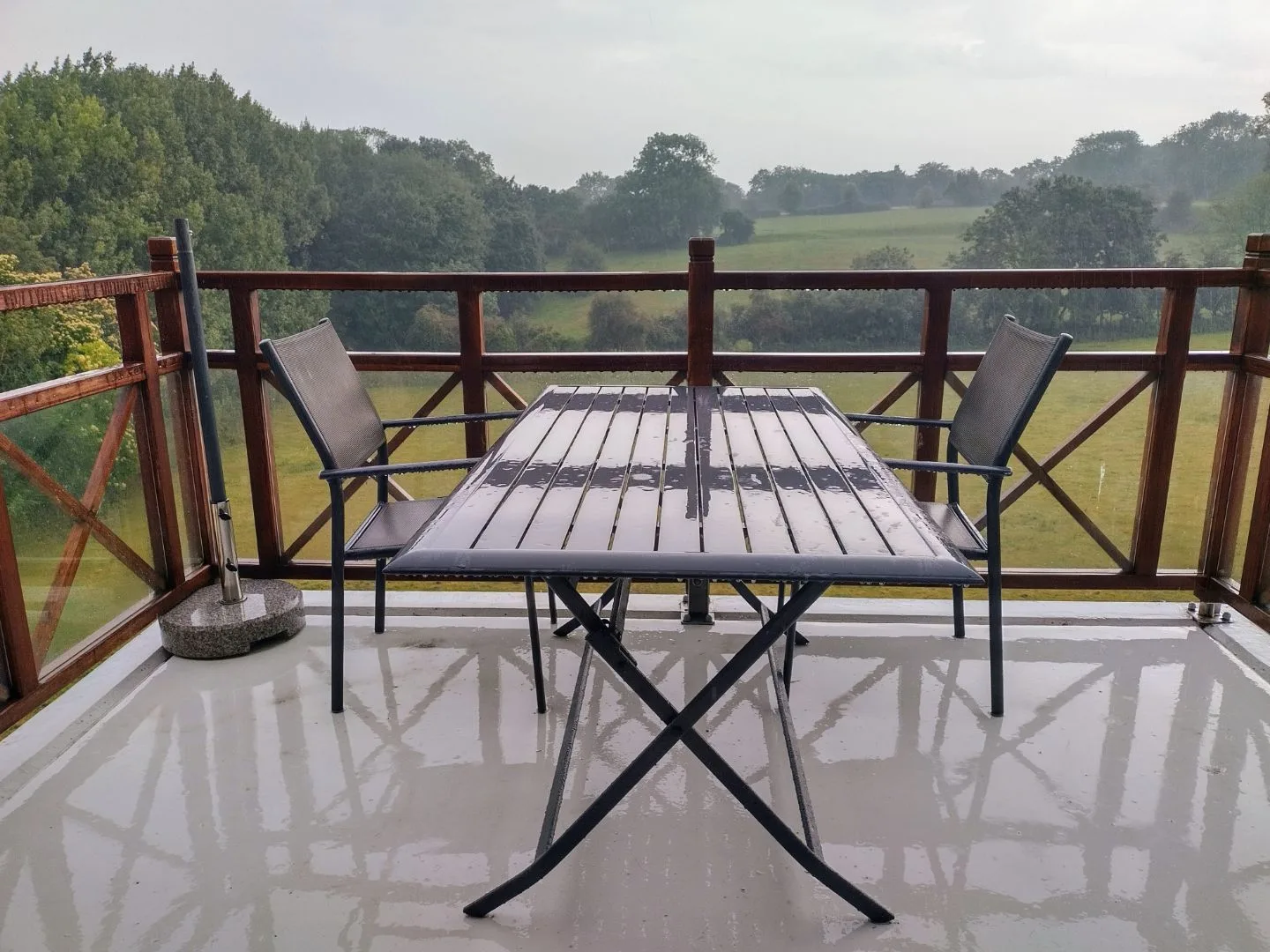 Ein regennasser Balkon mit zwei Stühlen und einem Tisch. Im Hintergrund eine Landschaft mit Wiesen und Bäumen