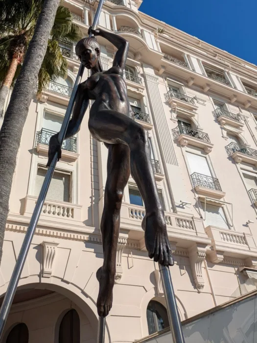 Das Bild zeigt eine Bronze-Skulptur einer nackten weiblichen Figur, die scheinbar an zwei hohen, schlanken Metallstangen balanciert. Im Hintergrund steht ein großes, hellbeiges Gebäude mit vielen Fenstern. Ein Palmenbaum ist teilweise links im Bild zu sehen. Der Himmel ist blau und sonnig. 