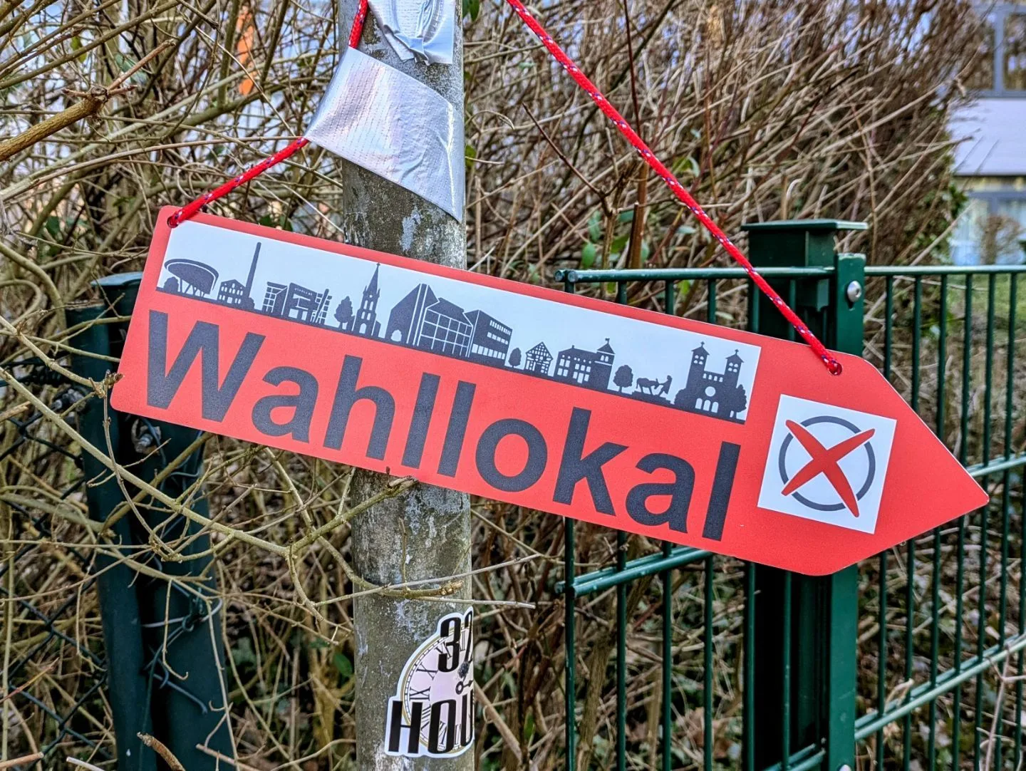 Das Bild zeigt ein rotes, pfeilförmiges Schild mit der Aufschrift "Wahllokal". Es hängt mit roter Schnur an einem Baum. Auf dem Schild ist eine Skyline abgebildet. Es dient als Wegweiser zu einem Wahllokal.