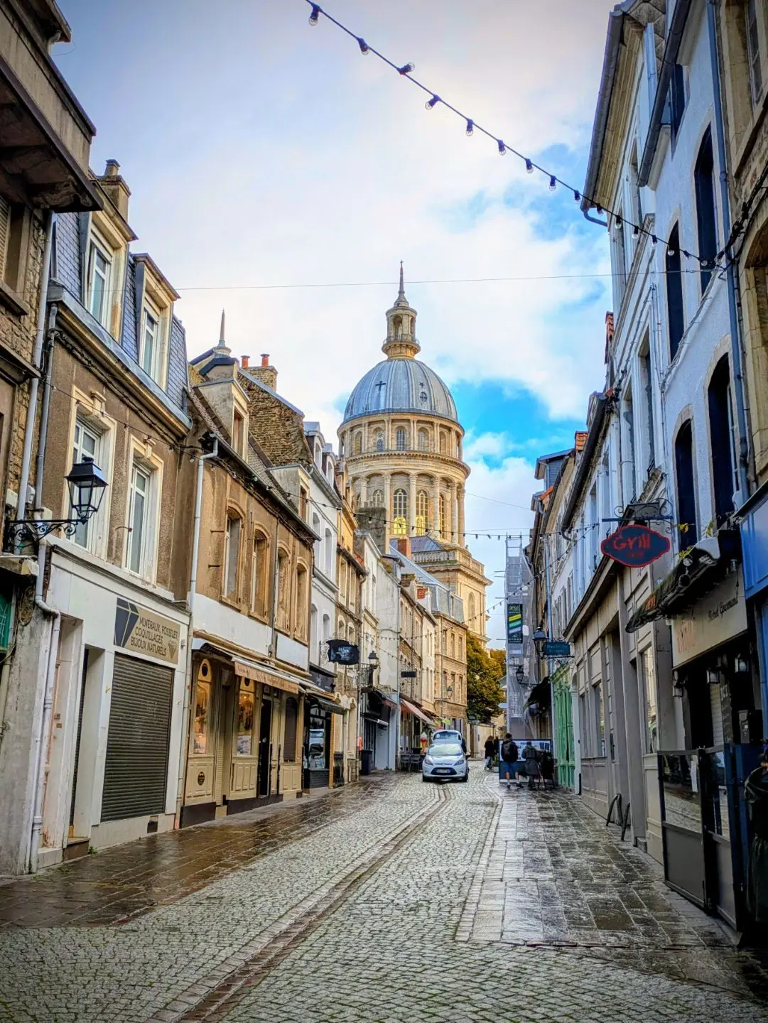 Pflasterstraße mit historischen Gebäuden und einer Kuppel im Hintergrund unter blauem Himmel mit Wolken.