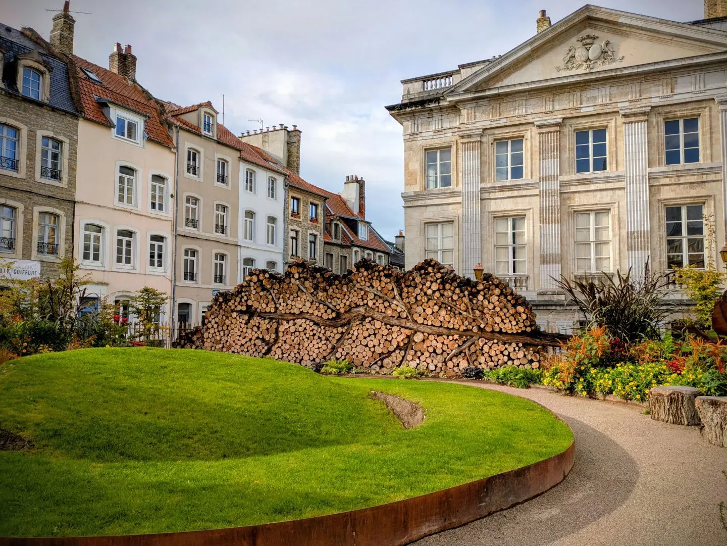 Rund geschwungener Weg und Rasenfläche vor einem großen Holzstapel in Form eines Blatts, umgeben von historischen mehrstöckigen Gebäuden.