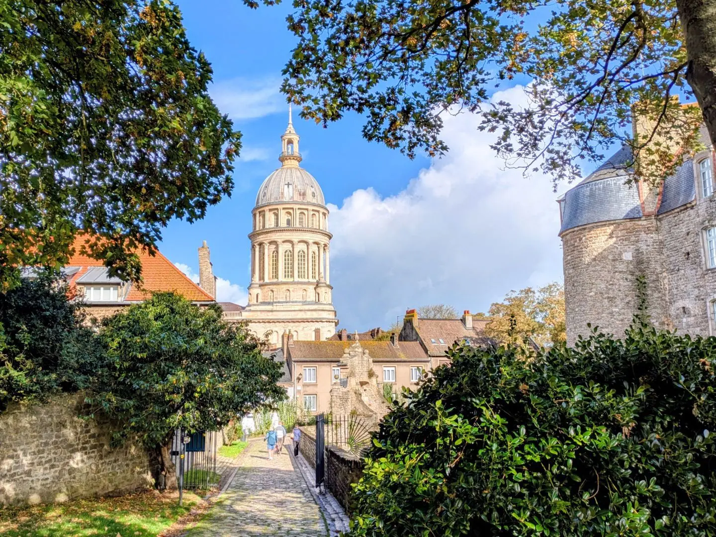 Blick auf einen historischen Turm hinter Bäumen und einem gepflasterten Weg mit zwei Personen, umgeben von alten Gebäuden unter blauem Himmel.