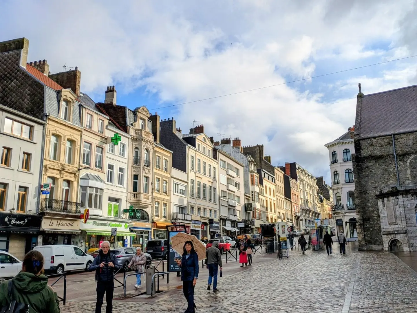 Straße mit Kopfsteinpflaster und mehrstöckigen Häusern mit Geschäften, darunter eine Apotheke mit grünem Kreuz, bei bewölktem Himmel.