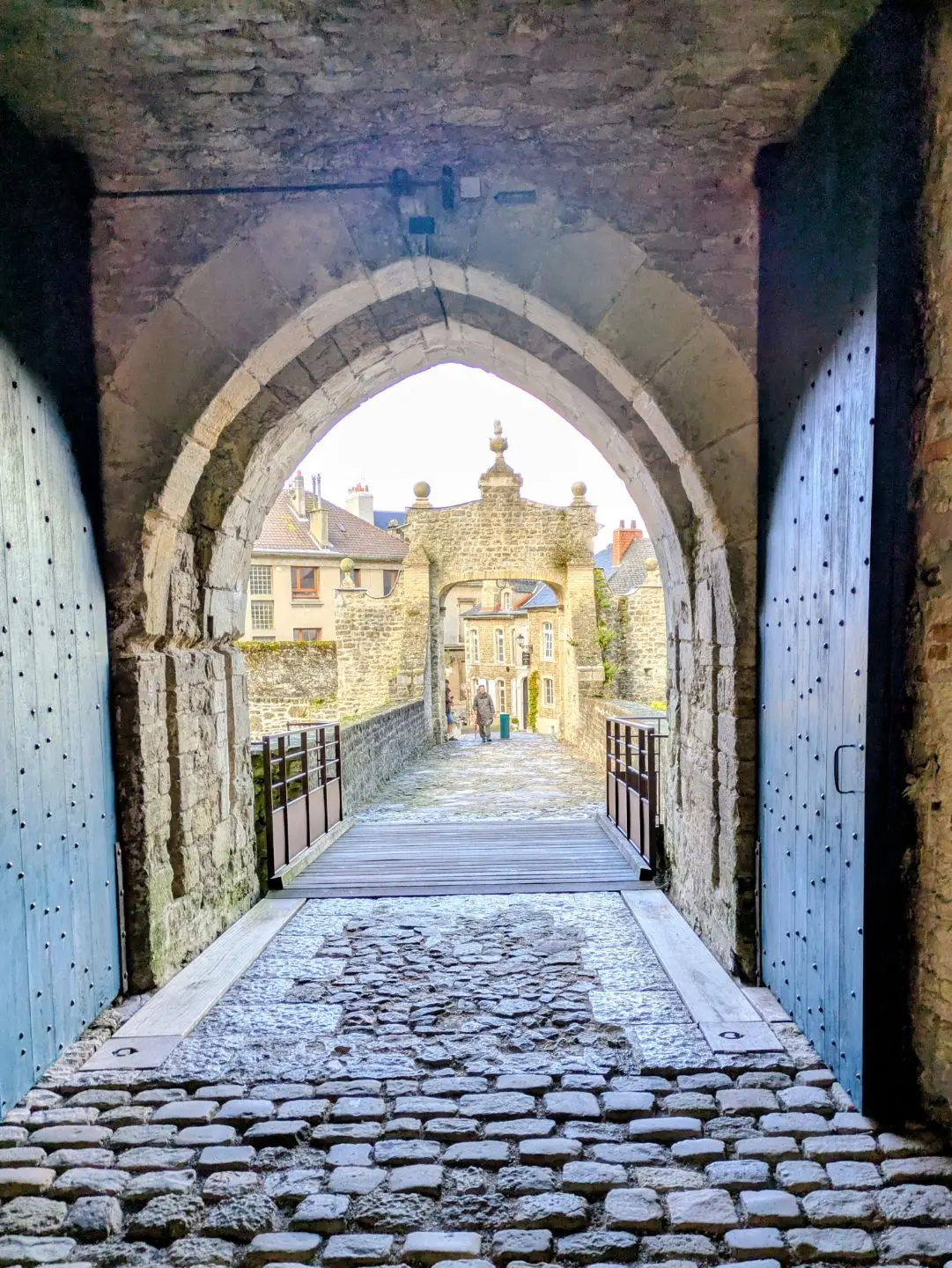 Blick durch ein steinernes Tor mit Holztüren auf eine gepflasterte Brücke und ein historisches Gebäude mit Zinnen und Türmchen.
