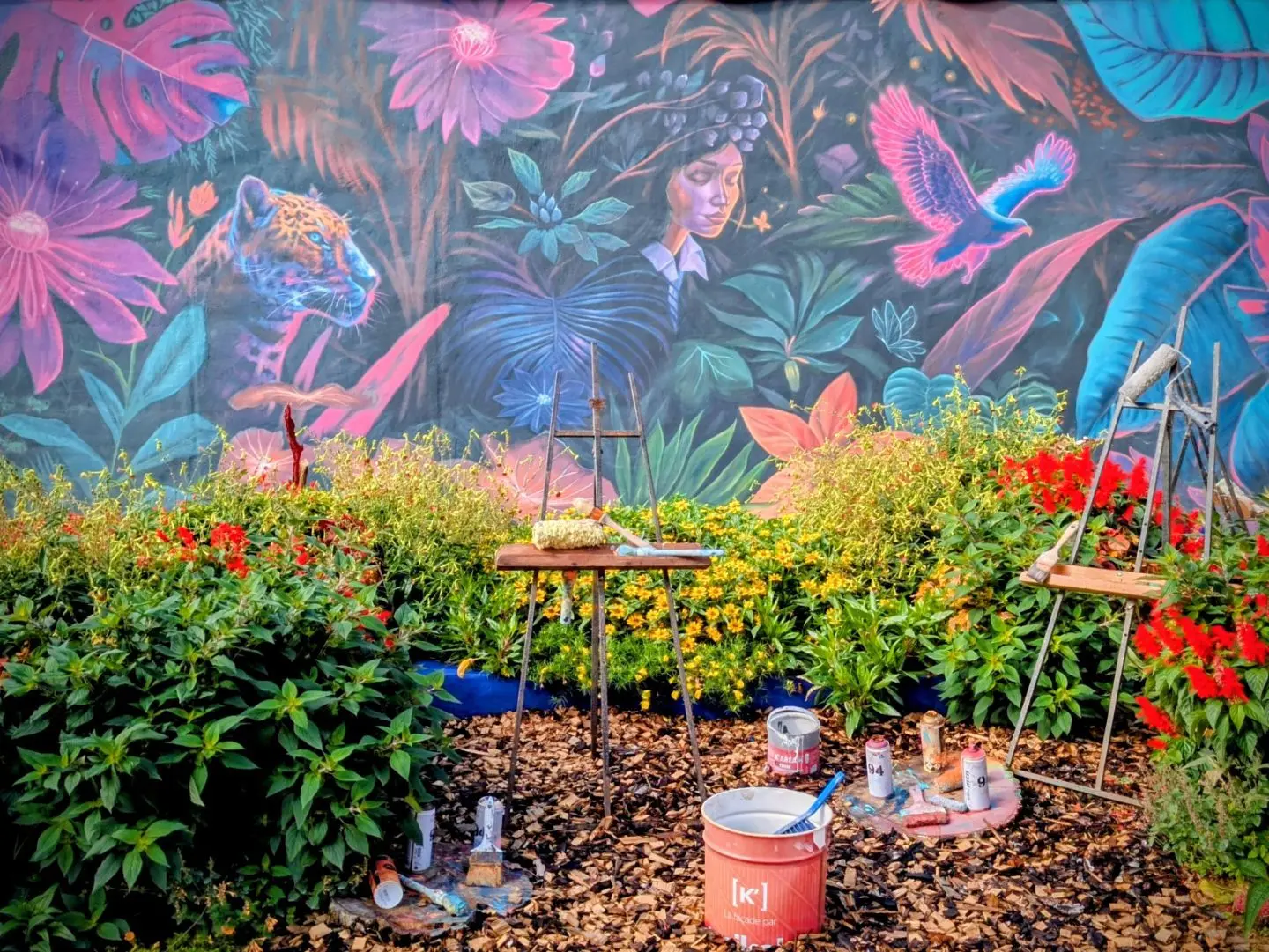Bunte Wandmalerei mit tropischen Pflanzen, einem Jaguar und einem Vogel, davor Garten mit Blumen, Staffelei und Malutensilien.
