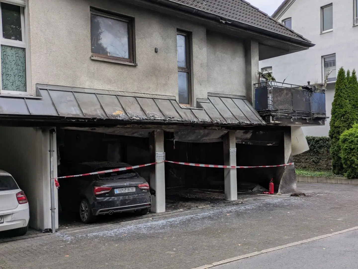 Dach eines Carports mit Brandschäden, Absperrband und zerbrochenem Glas auf dem Boden, zwei geparkte Autos darunter.