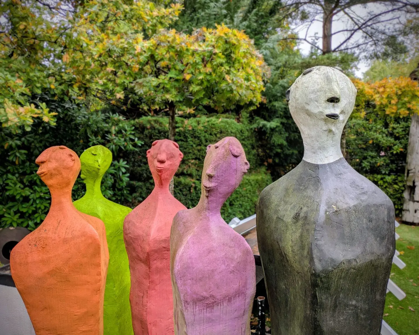 Fünf abstrakte, farbige Skulpturen mit menschlichen Köpfen in einem Garten, Farben: orange, grün, rot, violett, grau.