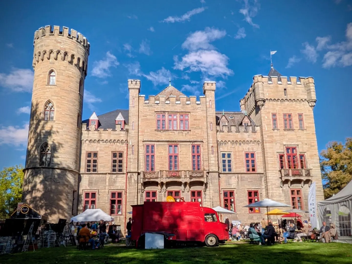 Historisches Schloss mit runden Türmen und roten Fenstern, davor ein roter Lieferwagen und Menschen bei einem Markt auf der Wiese.