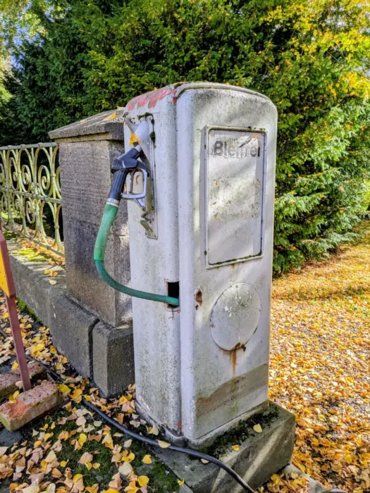Alte, weiße Zapfsäule mit grünem Schlauch und Zapfpistole neben einem Steinzaun, umgeben von Herbstlaub und Bäumen.