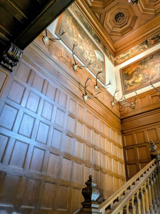 Holzvertäfelte Wand mit mehreren Geweihen und Deckenmalereien in einem Treppenhaus mit Holzgeländer.