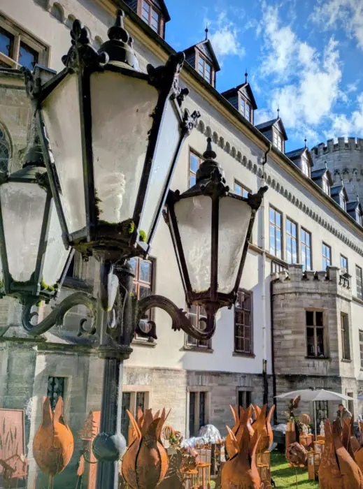 Zwei antike Straßenlaternen aus Metall vor einem historischen Gebäude mit Erkern und Dachgauben bei blauem Himmel.
