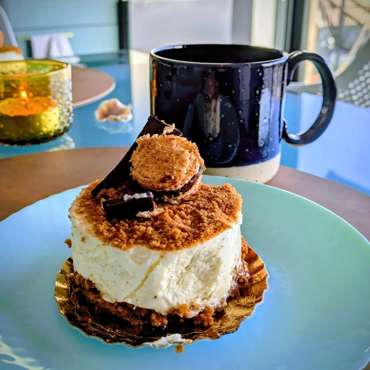 Kleiner runder Kuchen mit Keks- und Schokoladendekor auf blauem Teller, im Hintergrund dunkelblaue Tasse.
