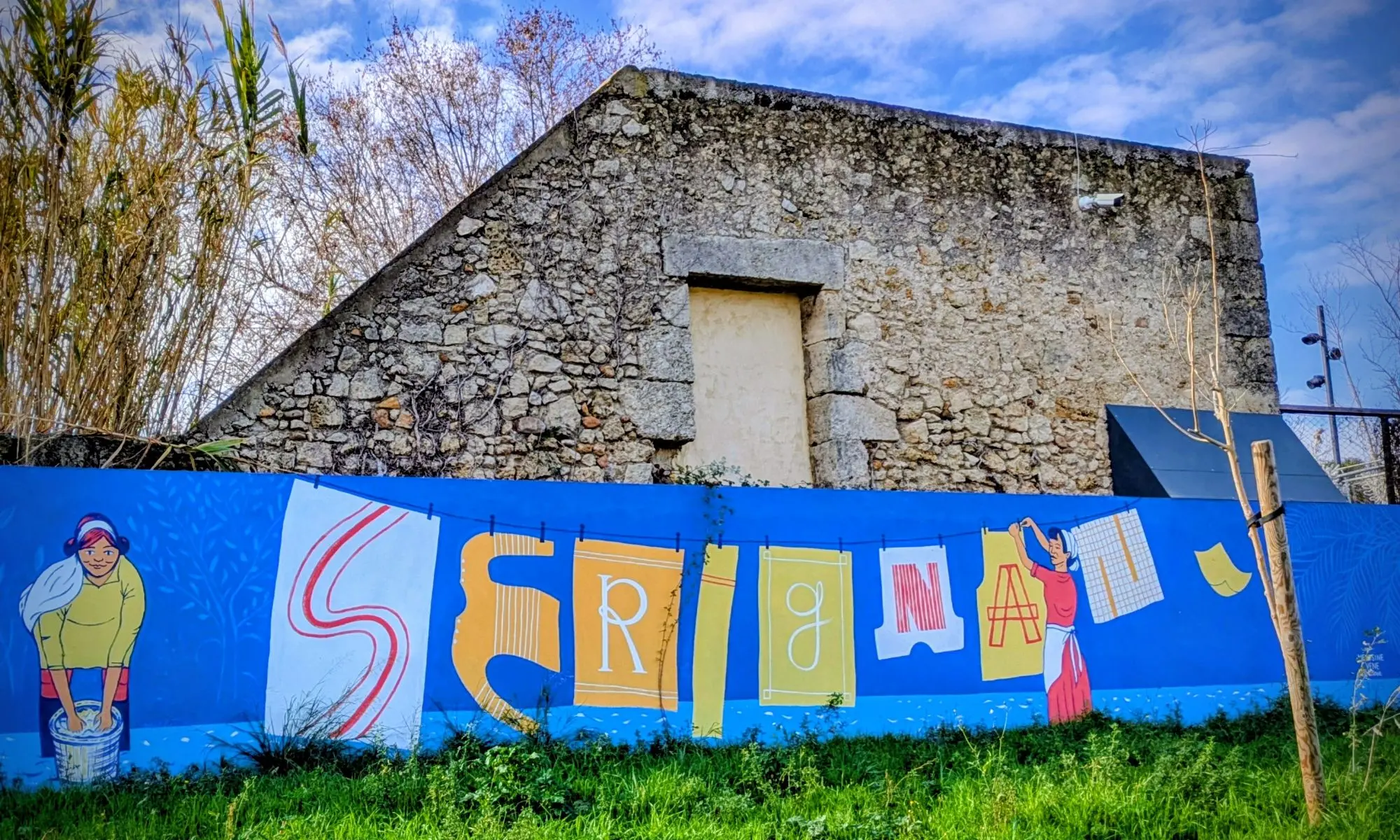 Bunte Wandmalerei mit Buchstaben auf blauem Hintergrund vor einer alten Steinmauer und Gras im Vordergrund