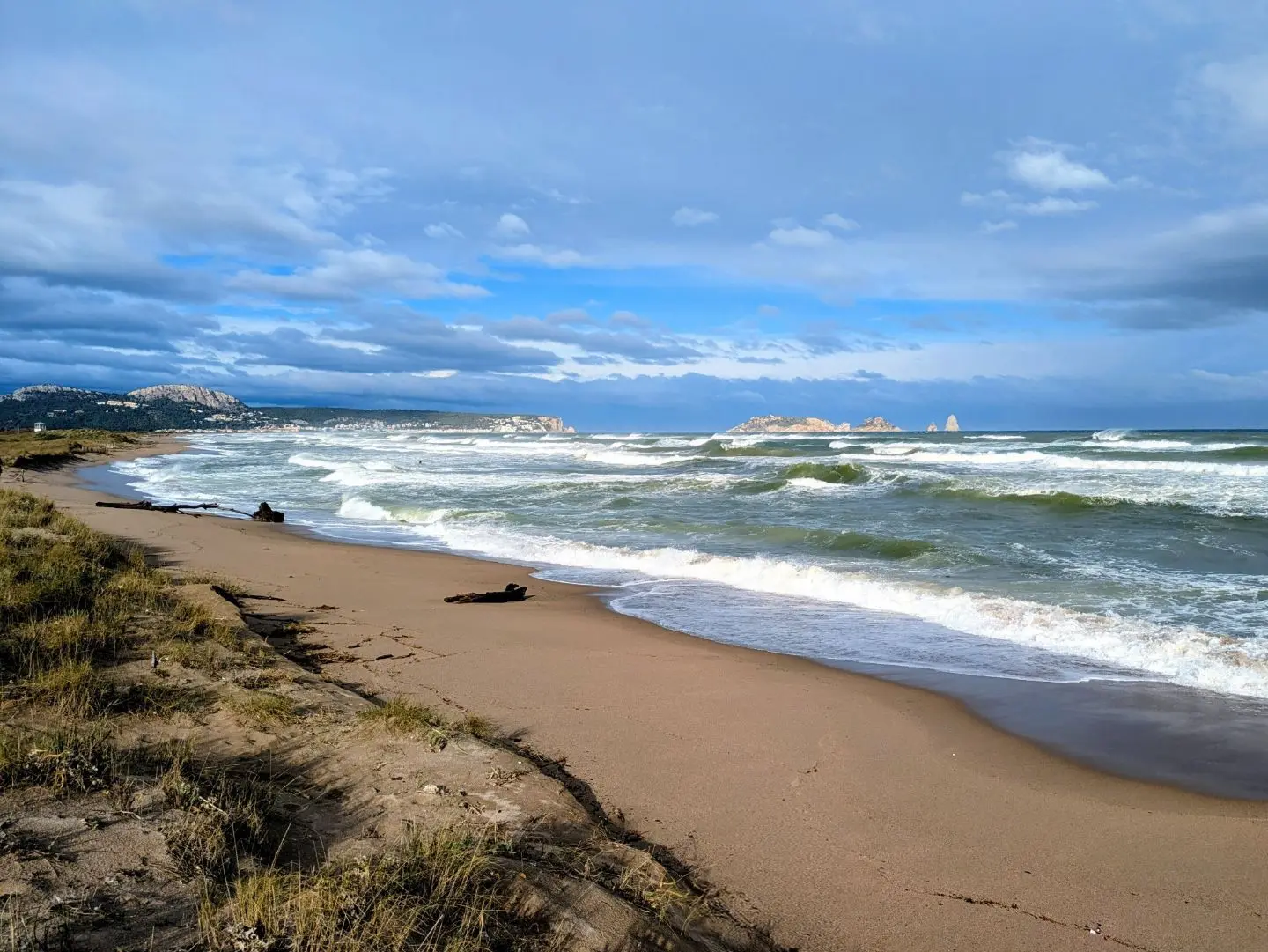 Strand mit Sand, Dünenbewuchs und Wellen unter bewölktem Himmel, im Hintergrund Felsen im Meer.