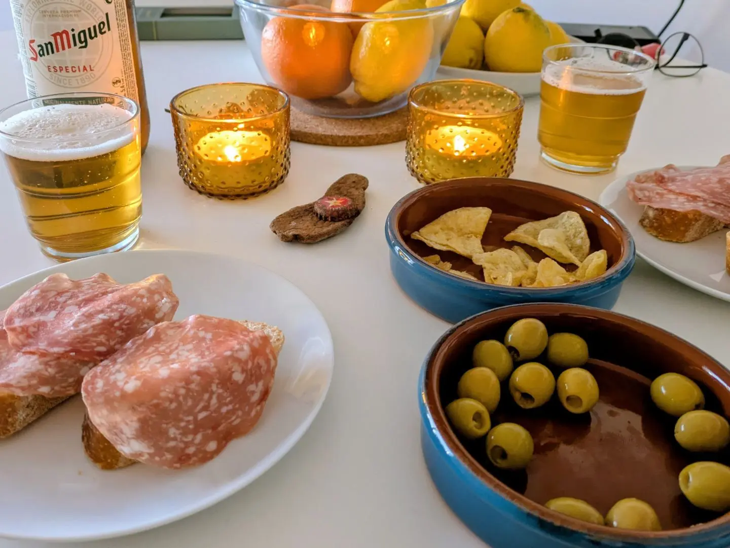 Tisch mit zwei Gläsern Bier, zwei Tellern mit Salami auf Brot, Schalen mit grünen Oliven und Chips, zwei brennenden Kerzen und einer Obstschale im Hintergrund.
