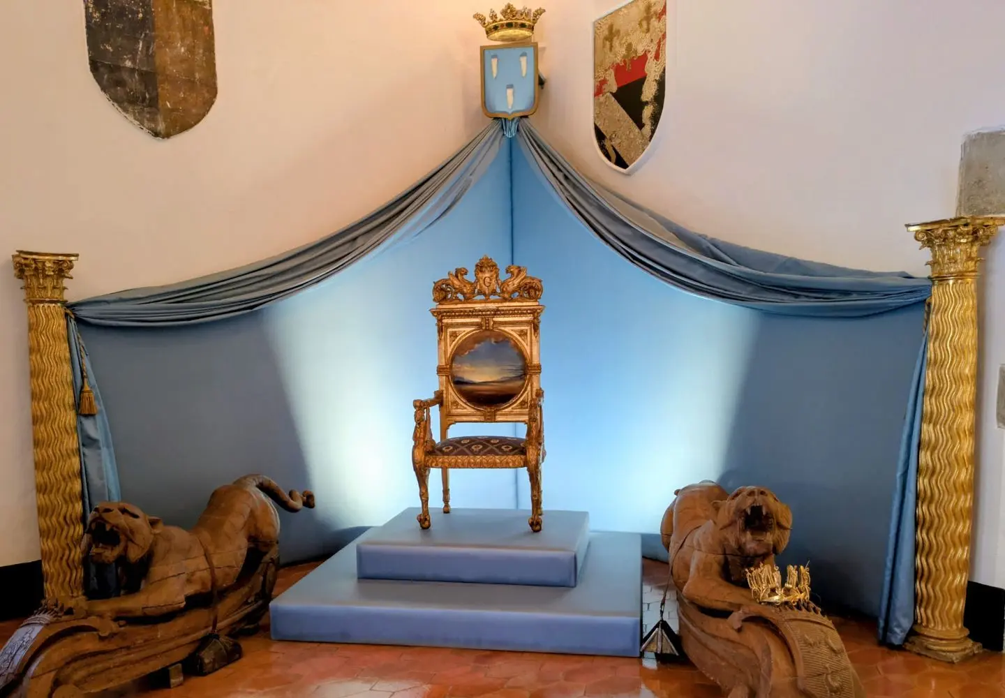 Prunkvoller Thron mit goldverzierten Armlehnen auf Podest, umgeben von zwei liegenden Löwenfiguren und zwei Säulen, im Hintergrund blaues Tuch mit Wappen und Krone
