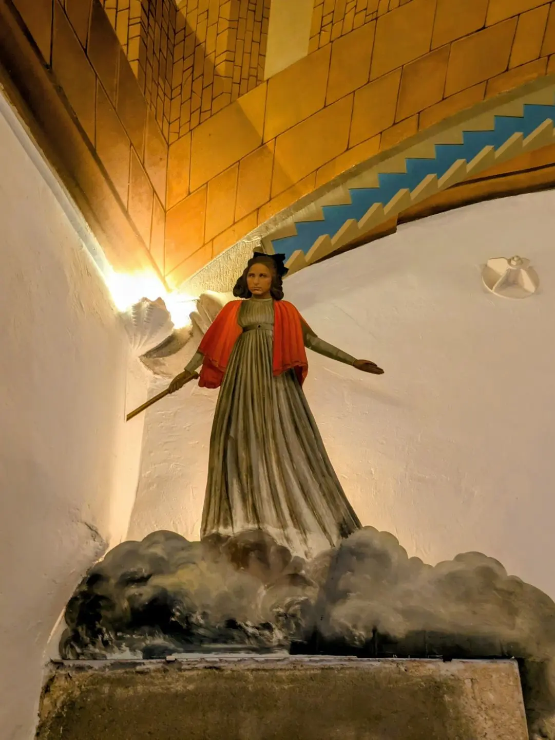 Statue einer Frau mit ausgebreiteten Armen, die ein rotes Tuch trägt und auf einer Wolkenbasis steht, vor einer gelb gekachelten Wand mit Treppenbogen