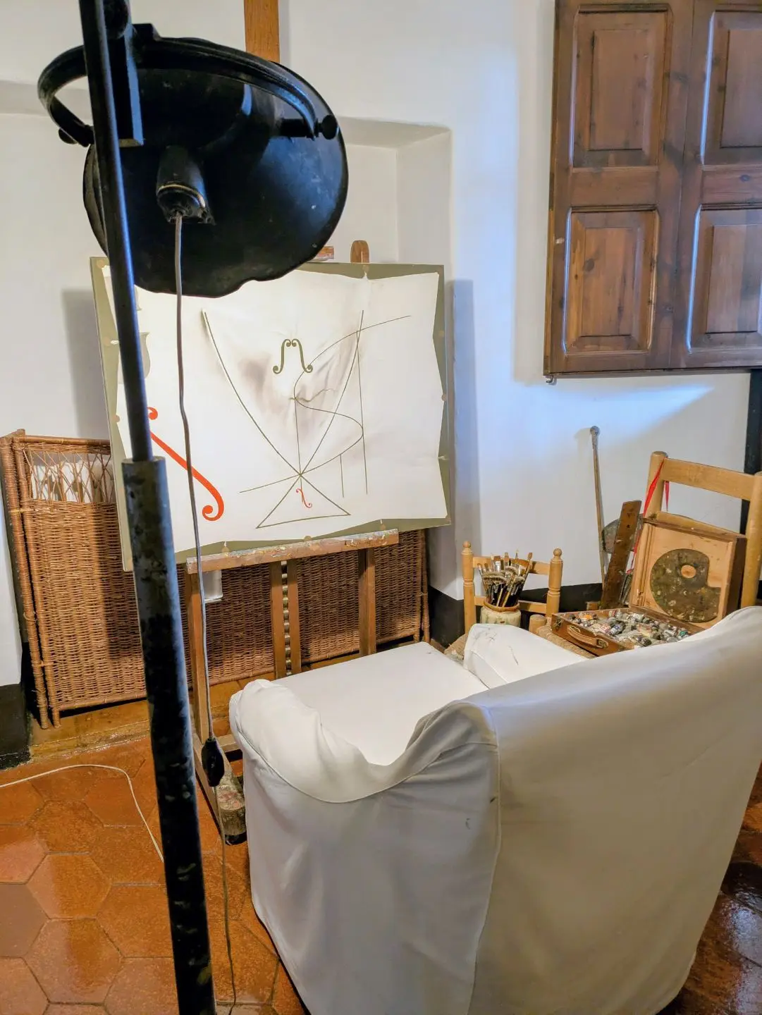 Wohnzimmer mit weiß bezogenem Sessel, Staffelei mit abstraktem Bild, Holzstuhl mit Kissen und einem großen schwarzen Stehlampen-Schirm