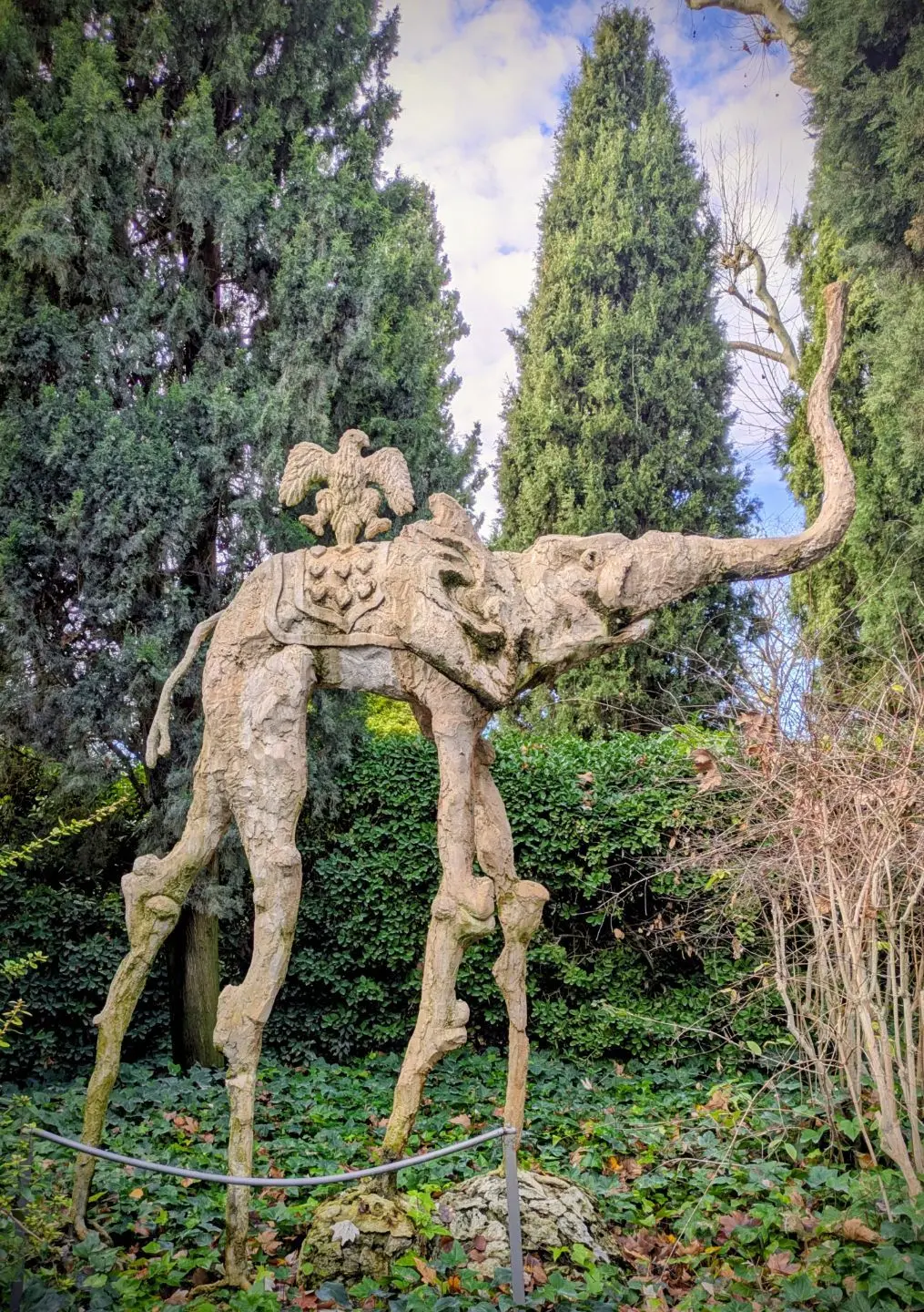 Skulptur eines Elefanten mit langen, dünnen Beinen und einem Adler auf dem Rücken, umgeben von Bäumen und Pflanzen