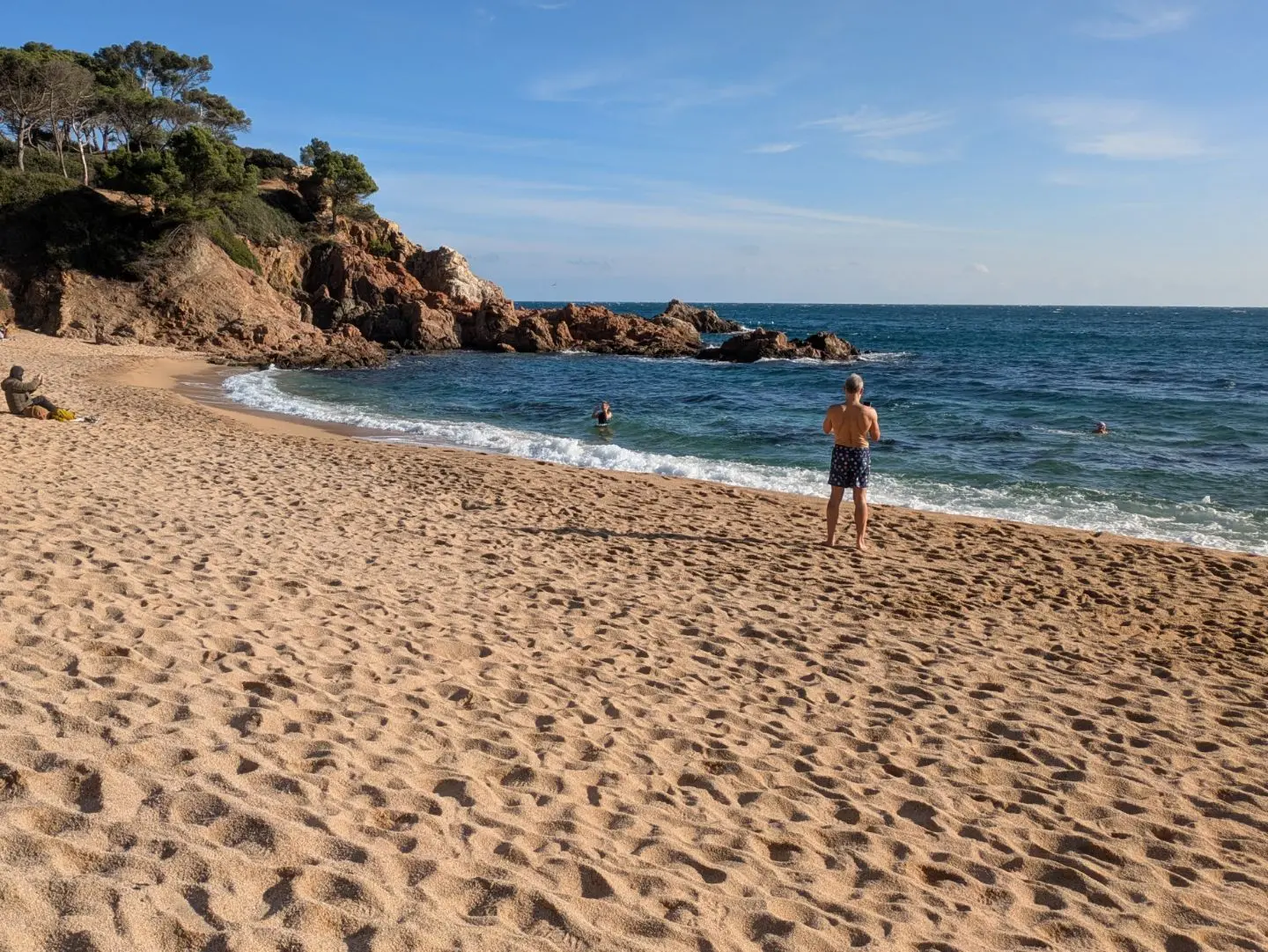 Strand mit Sand, Felsen und mehreren Personen, von denen eine am Ufer steht und andere im Wasser schwimmen