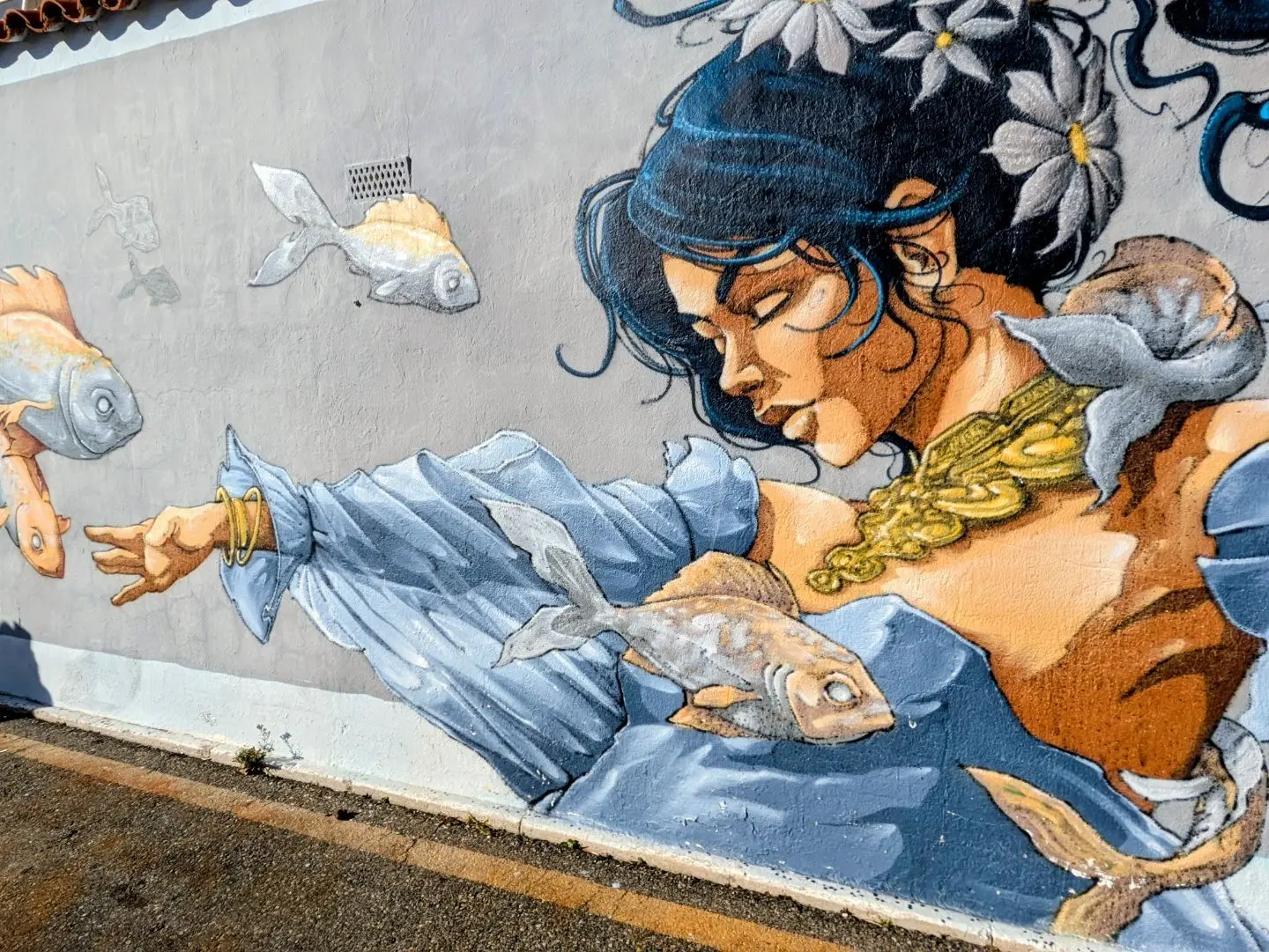 Wandgemälde einer Frau mit blauer Kleidung und goldener Halskette, umgeben von mehreren Fischen.