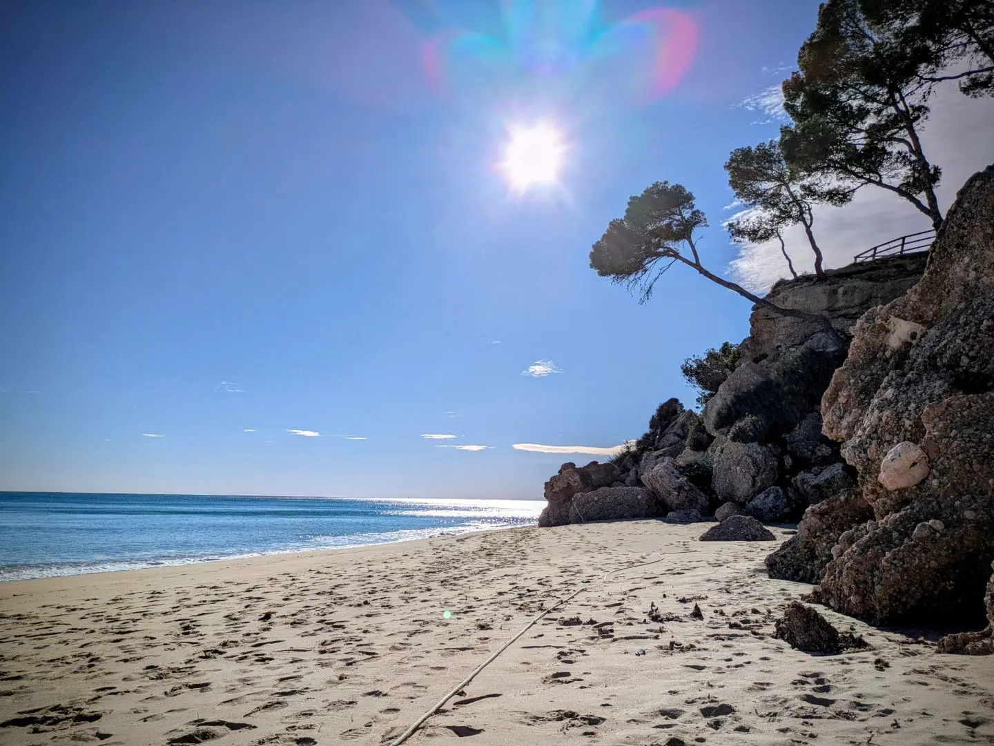 Strand mit hellem Sand, Felsen und vereinzelten Bäumen rechts, ruhiges Meer links und Sonne am klaren Himmel.