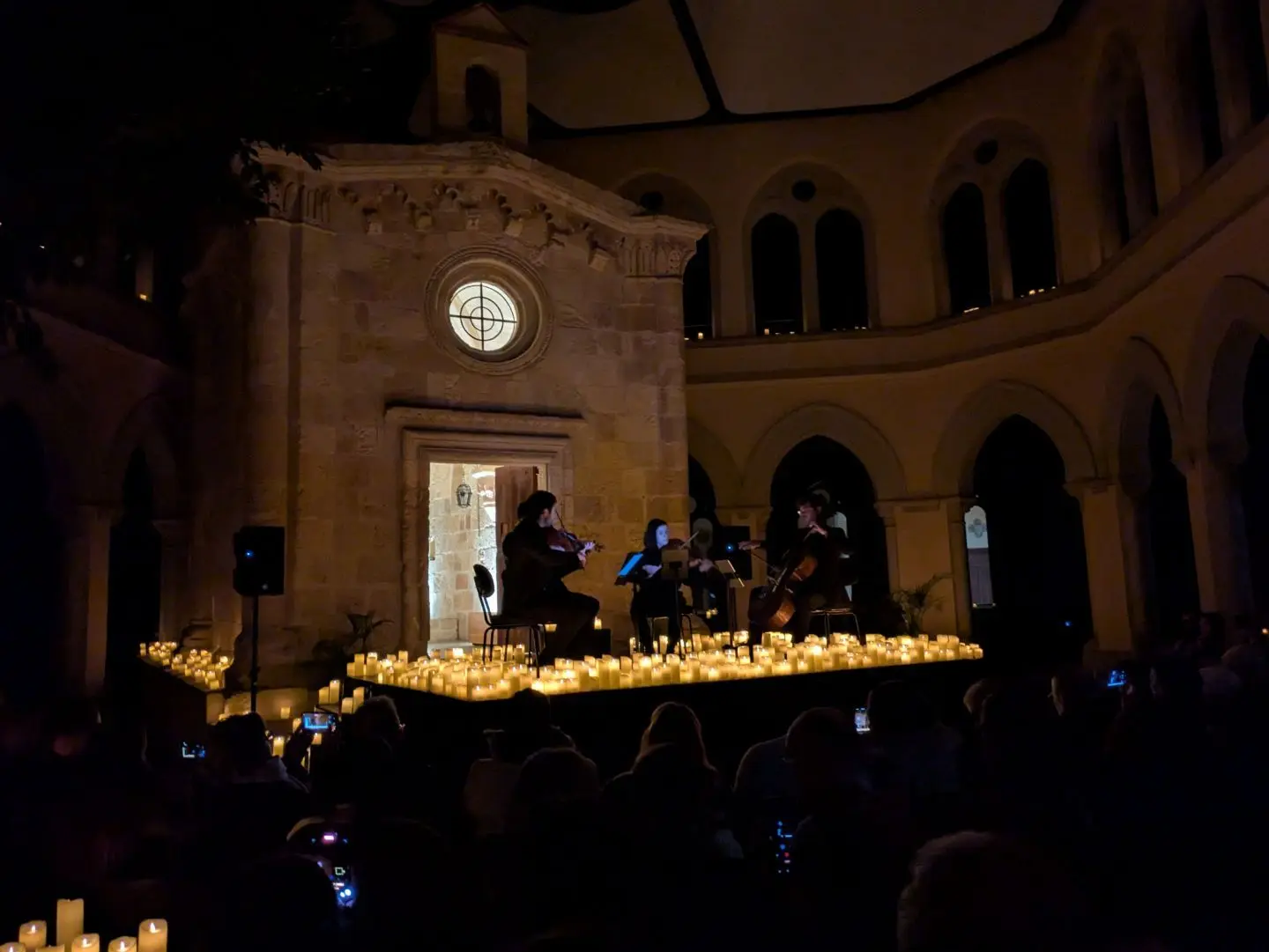 Vier Musiker spielen in einer dunklen Kirche auf einem von vielen Kerzen umgebenen Podium, Publikum im Vordergrund mit Handys.