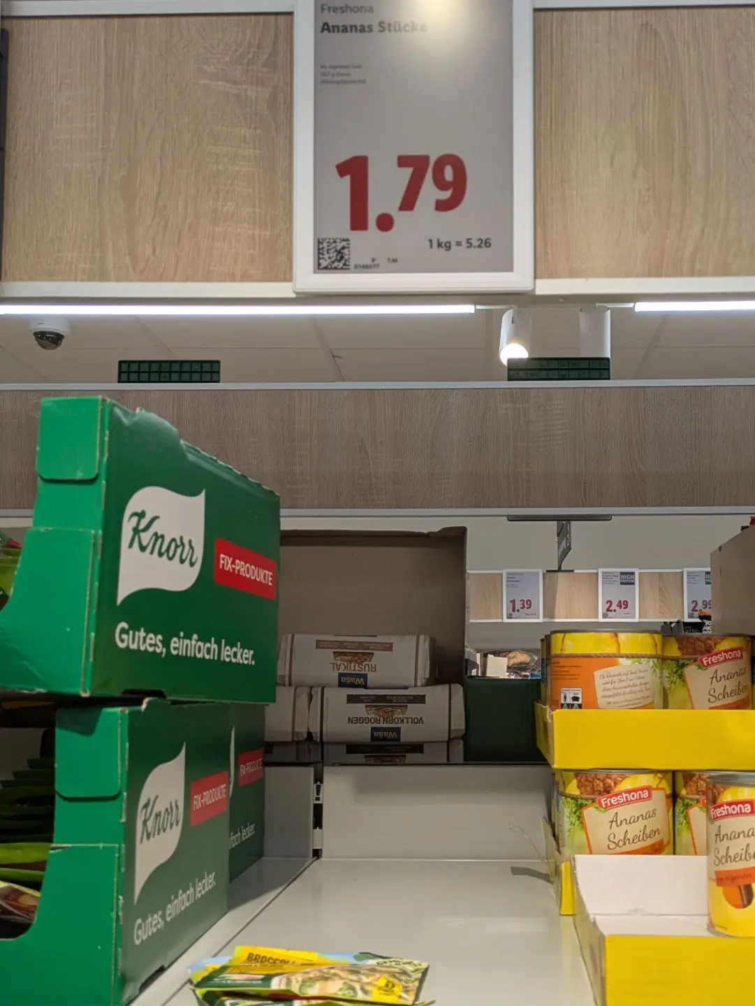 Regal mit grünen Knorr-Produkten links und gelben Freshona-Ananas-Scheiben rechts in einem Supermarkt mit Preisschild '1.79'