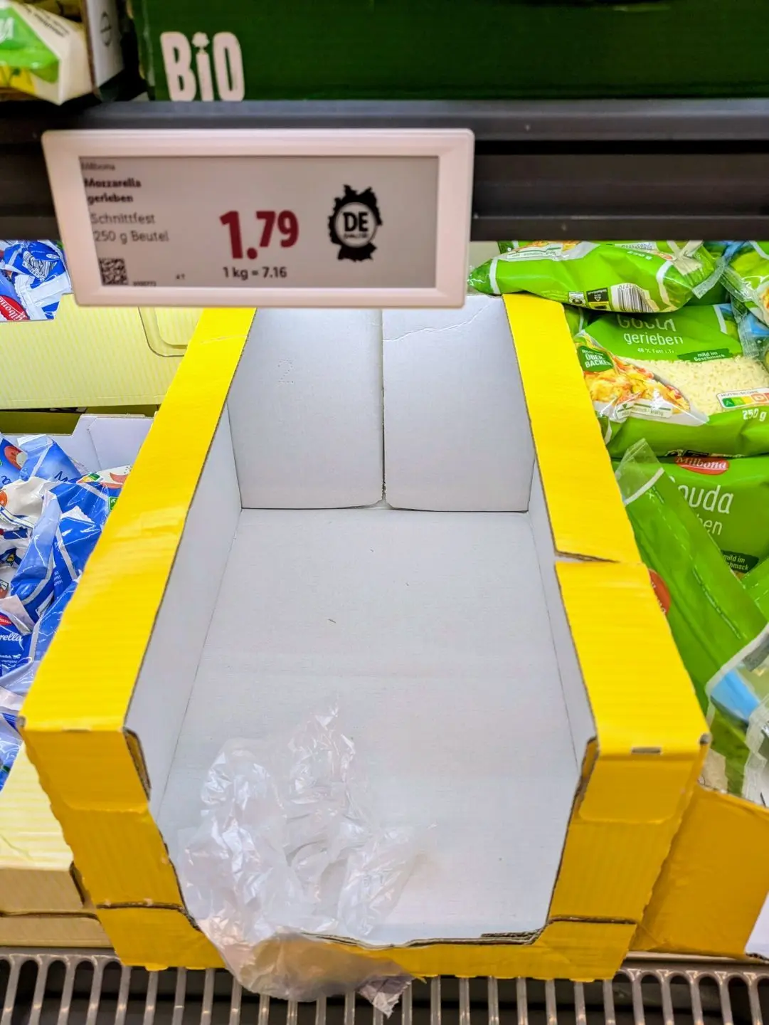 Leeres gelb umrandetes Pappfach in einem Kühlregal mit Preisschild für Mozzarella-Schnittkäse