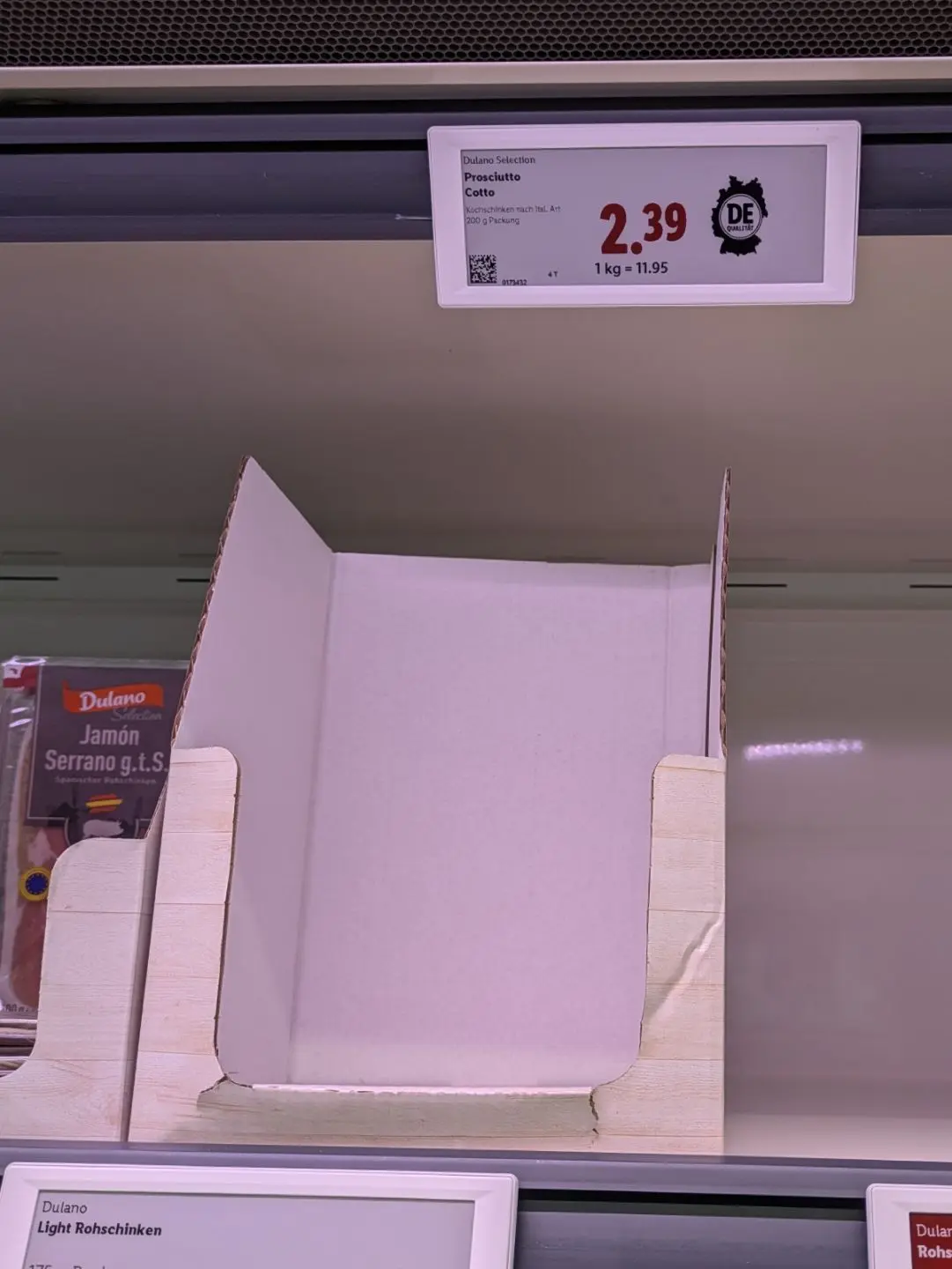 Leeres weißes Pappregalfach mit Preisschild für Prosciutto und links daneben verpackte Schinkenprodukte