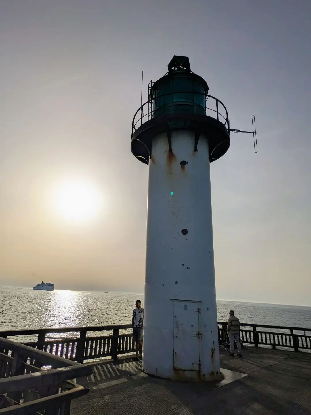 Leuchtturm auf einem Steg mit zwei Personen und einem Schiff auf dem Meer im Hintergrund bei Sonnenlicht