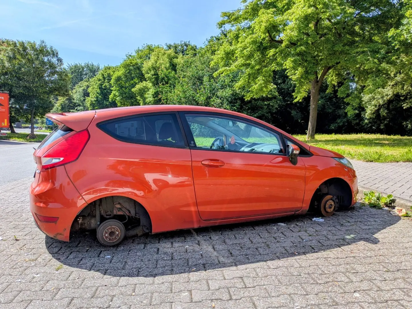 Oranger Kleinwagen steht auf gepflastertem Boden ohne Reifen, nur mit Bremsscheiben sichtbar