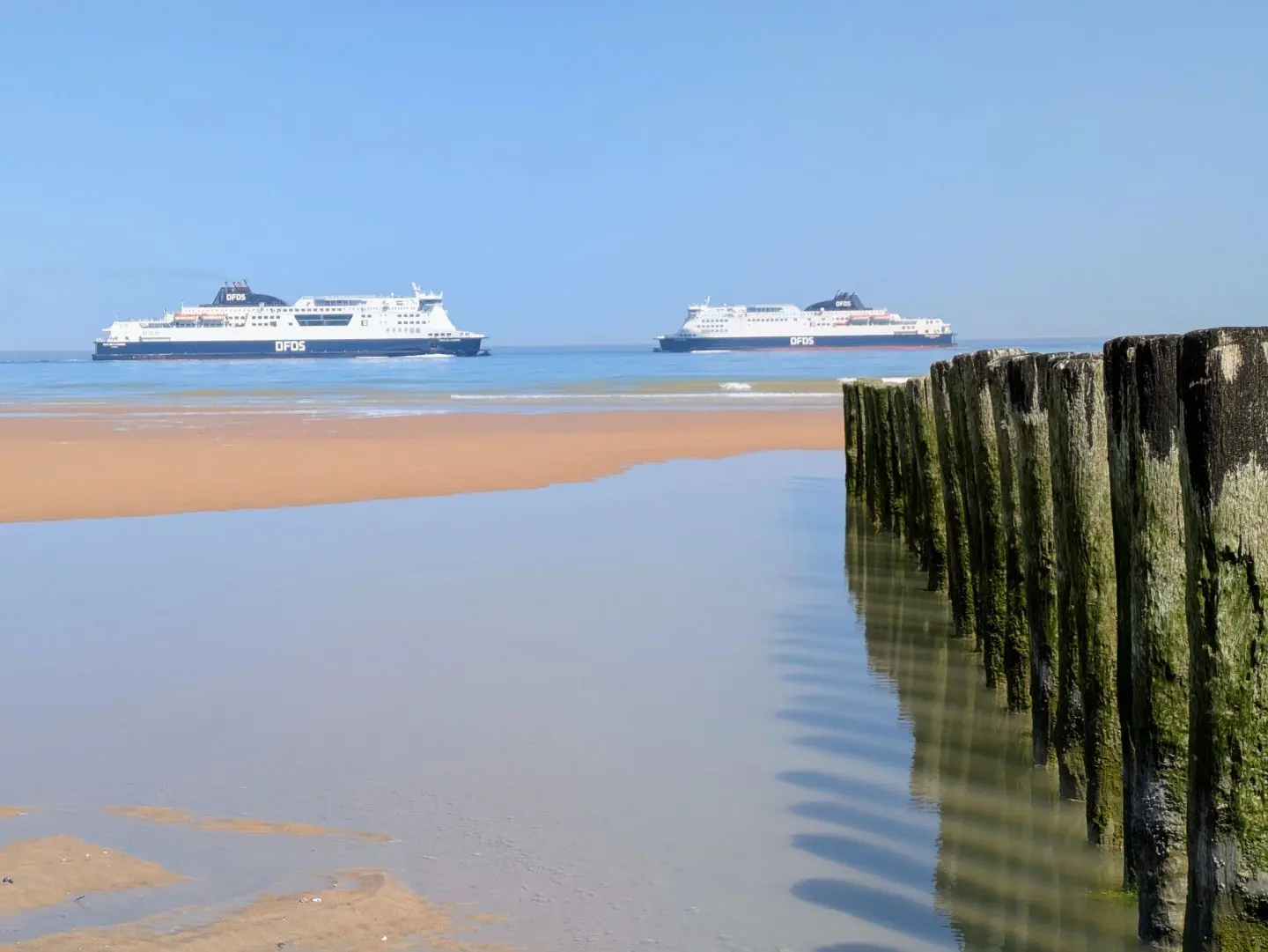 Zwei große Fähren auf ruhigem Meer vor einem Sandstrand mit einer Reihe von Holzpfählen im Wasser im Vordergrund